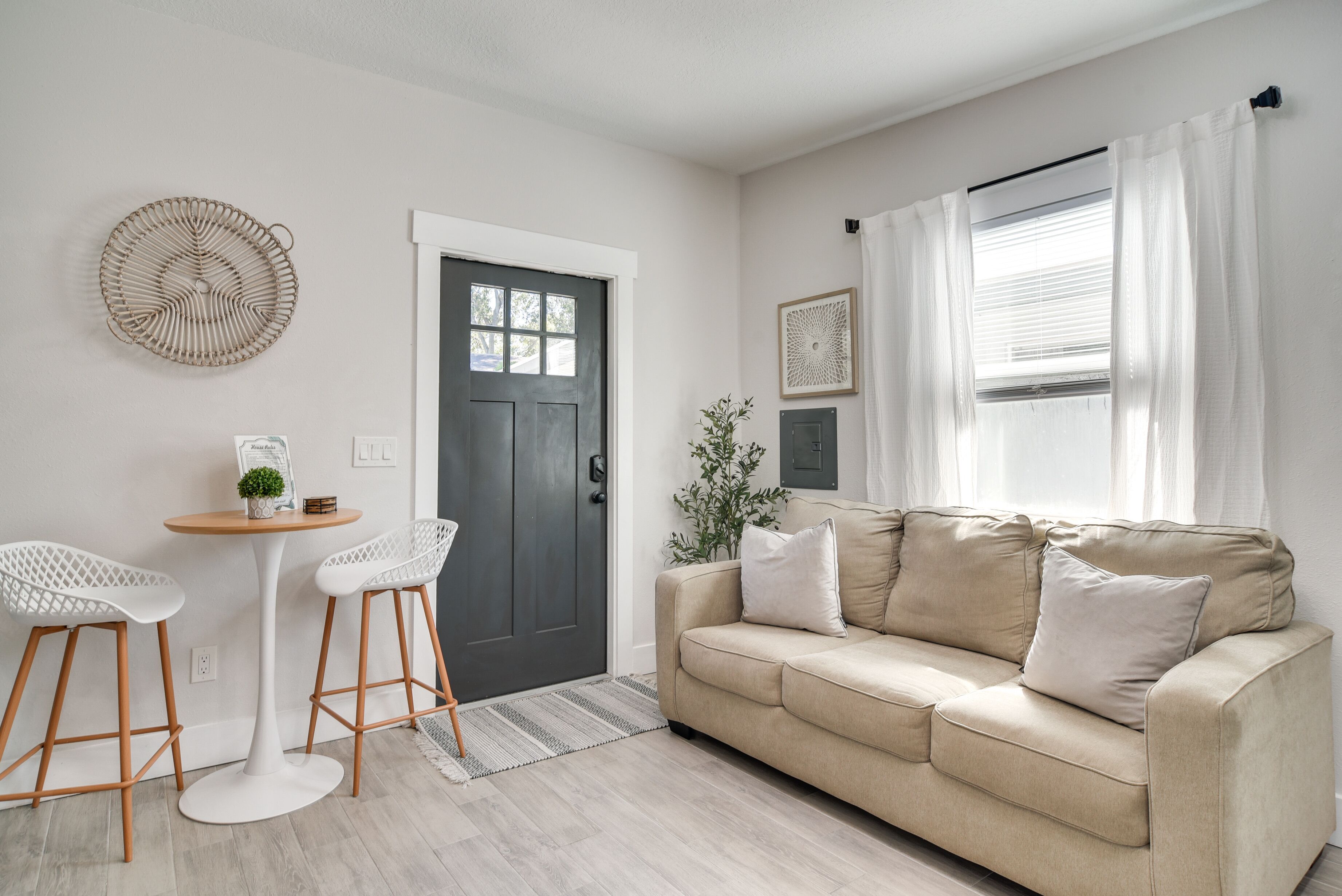 A living room with a beige sofa, two white chairs, a small round table, a dark door, and a window with white curtains.