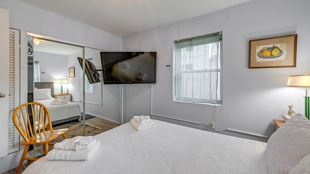 A hotel room with a large bed, a TV mounted on the wall, a wooden chair, and a painting on the wall. - Pet-friendly Great Neck Home ~ 1 Mi to Kings Point (Great Neck)