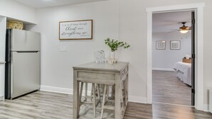 Une cuisine avec un réfrigérateur blanc, une table en bois ornée d’un vase et d’une citation encadrée, et un couloir menant à une chambre à coucher équipée d’un lit et d’un ventilateur de plafond.