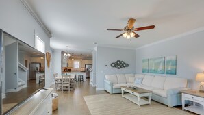 Un salon spacieux avec un canapé bleu clair, une table basse et un téléviseur. On y trouve également un ventilateur de plafond, une cuisine à l’arrière-plan et un escalier menant à un autre niveau.