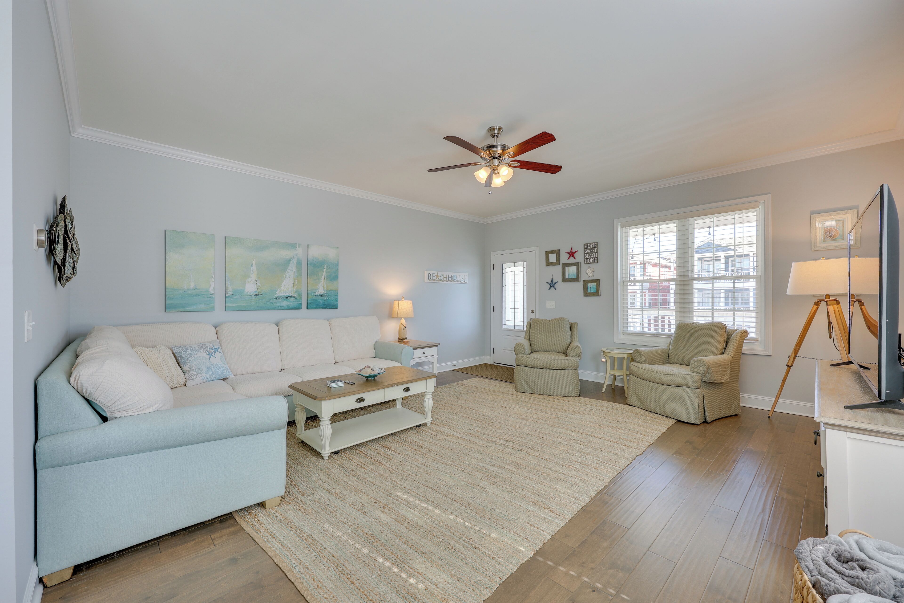 Ein Wohnzimmer mit einem hellblauen Sofa, einem Holztisch, einem beigefarbenen Sessel und einem weißen Schrank. Es gibt einen Deckenventilator, ein Fenster mit Jalousien und eine Lampe auf einem Beistelltisch.