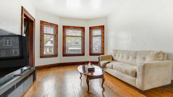 A living room with a flat-screen TV, a beige sofa, a wooden coffee table, and three windows with wooden frames.