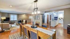 Een ruime woonkamer met een houten eettafel, stoelen en een moderne kroonluchter. Achtergrond is een keuken met witte kasten en een magnetron.