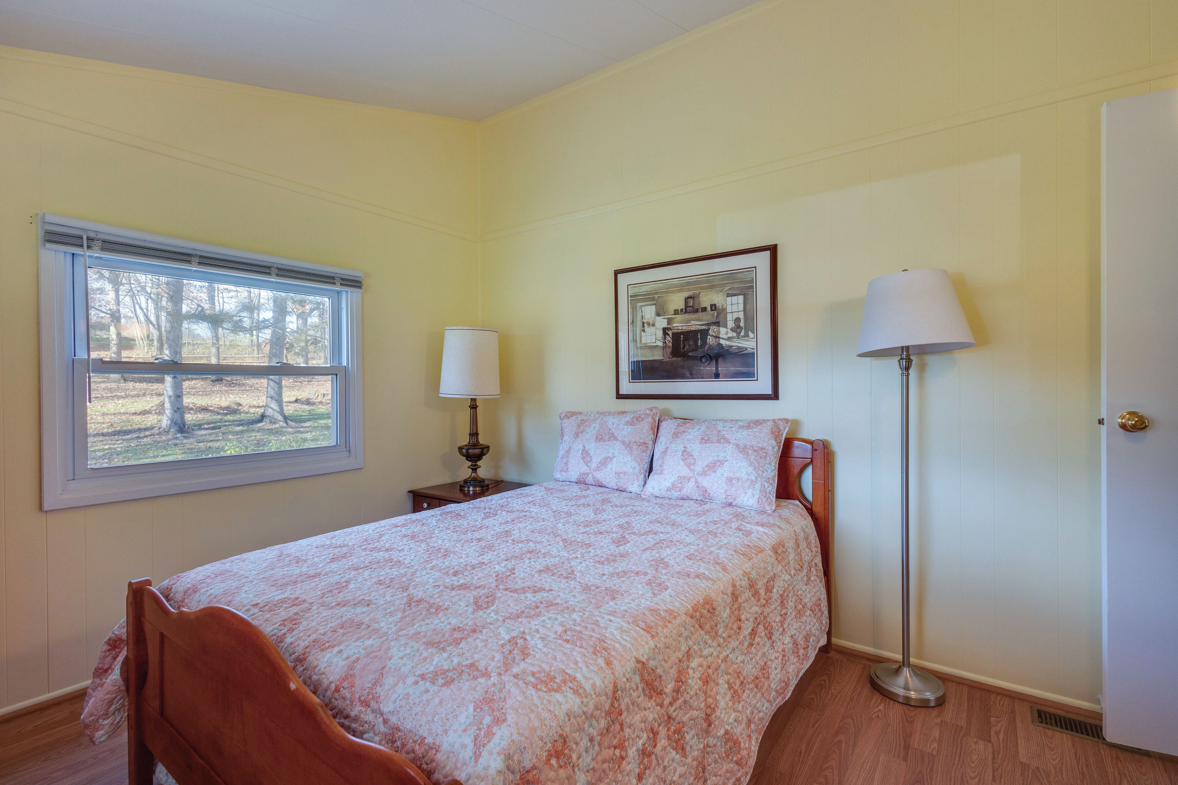 A bedroom with a bed, a window, a bedside table with a lamp, a picture on the wall, and a floor lamp.