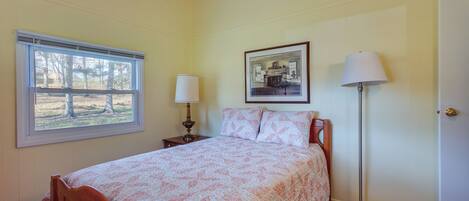 A bedroom with a bed, a window, a bedside table with a lamp, a picture on the wall, and a floor lamp.