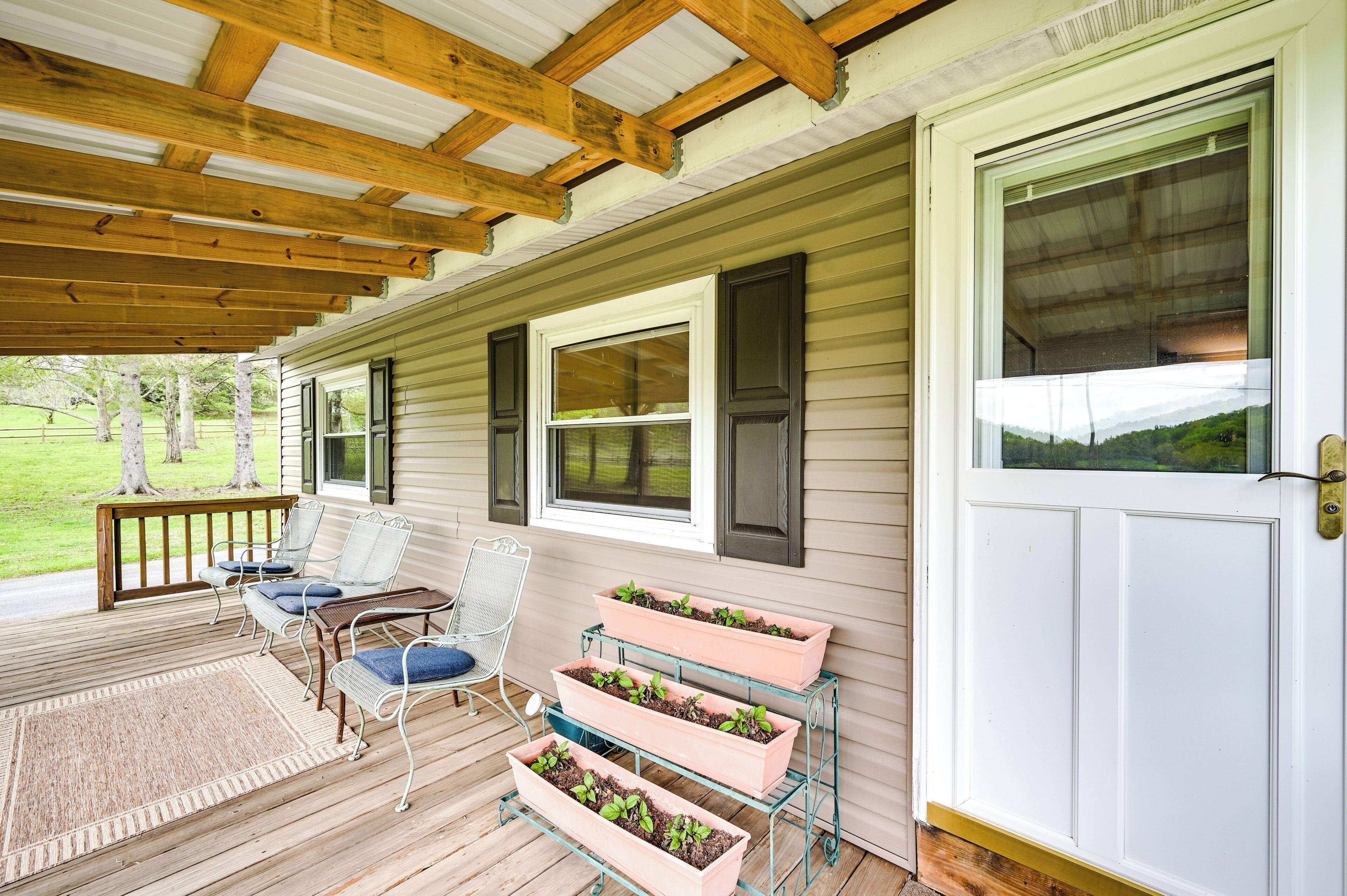 A covered porch with wooden beams, a white door, and a window with shutters. There are three metal planters with plants on the porch.
