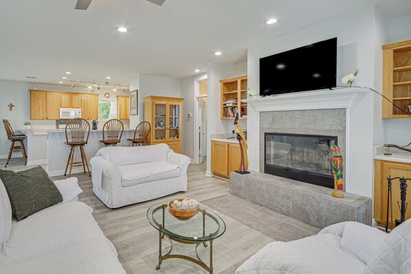 A modern living room with a fireplace, a television mounted above it, and a kitchen area in the background. - Waveland Vacation Rental - Walk to the Beach! (Waveland)