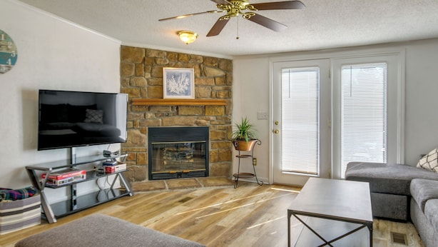 A living room with a stone fireplace, a flat-screen TV, a ceiling fan, and a large window with blinds. - Prosperity Vacation Rental ~ 1 Mi to Lake Murray! (Prosperity)