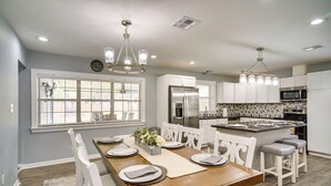 A modern kitchen with white cabinetry, stainless steel appliances, and a dining area with a wooden table and white chairs. - Lake Charles Home w/ Gas Grill & Fenced-in Yard! (Lake Charles)