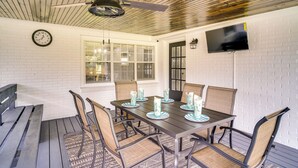 A dining area with a table set for six, surrounded by wicker chairs, a ceiling fan, a clock, and a TV mounted on the wall. - Lake Charles Home w/ Gas Grill & Fenced-in Yard! (Lake Charles)