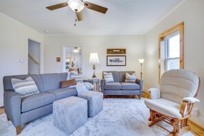 A living room with a grey sofa, ottoman, and a rocking chair. There is a ceiling fan, a window with a wooden frame, and a mirror on the wall.