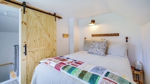 A bedroom with a wooden door, a quilted bedspread, a wall-mounted light, and a bedside table with a remote control. - Lovely Rural Hixton Farmhouse w/ Porch & Fire Pit! (Hixton)