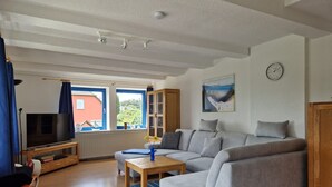 A living room with a grey sofa, a wooden coffee table, and a television on a stand. There is a window with blue curtains, a bookshelf, and a painting on the wall.