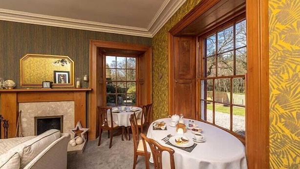 A dining area with a fireplace, wooden furniture, and a view through large windows. - Maxwelston Farmhouse (Girvan)