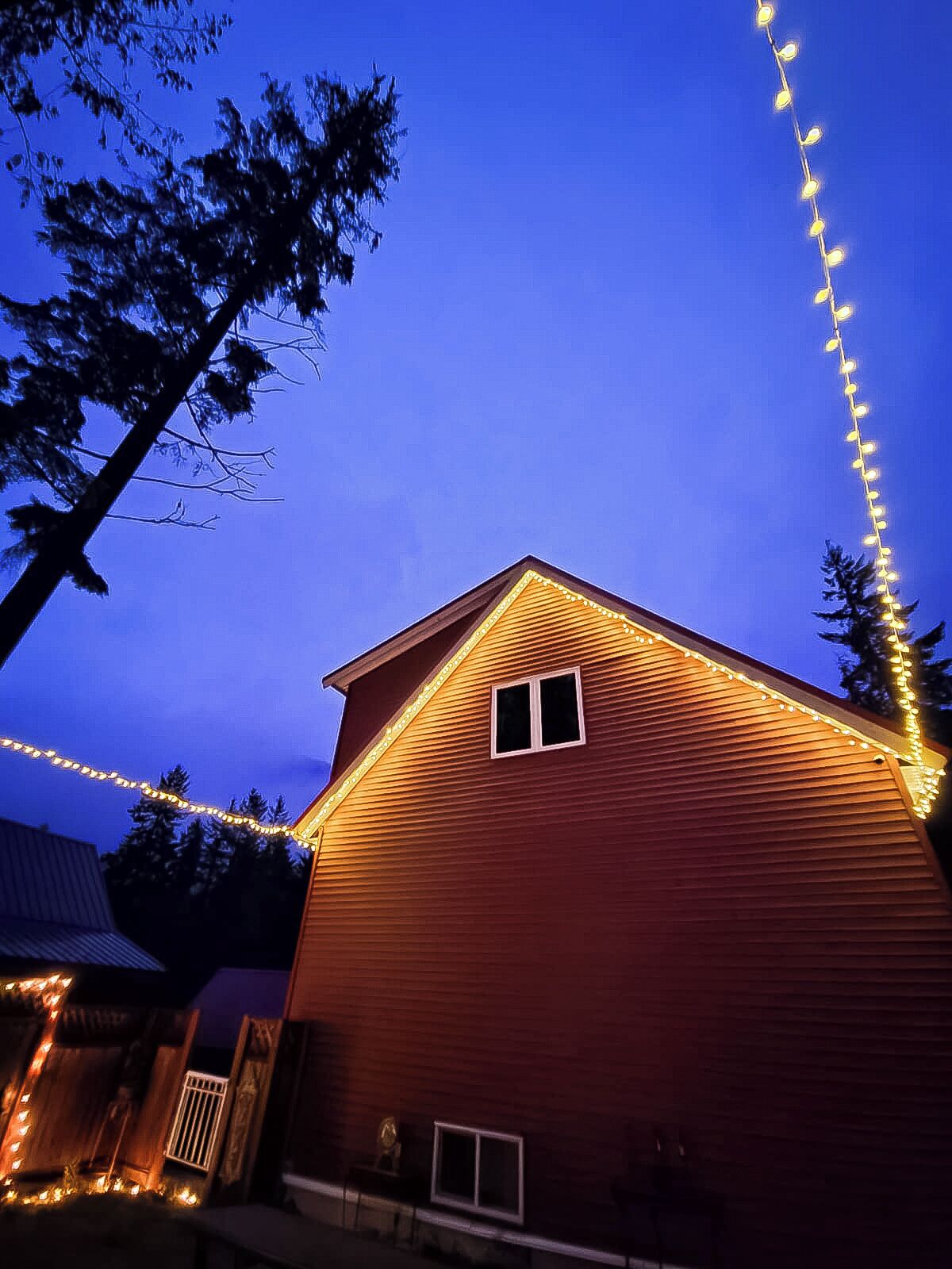 Una casa de madera con techo a dos aguas y una pequeña ventana, rodeada de árboles y luces colgantes.