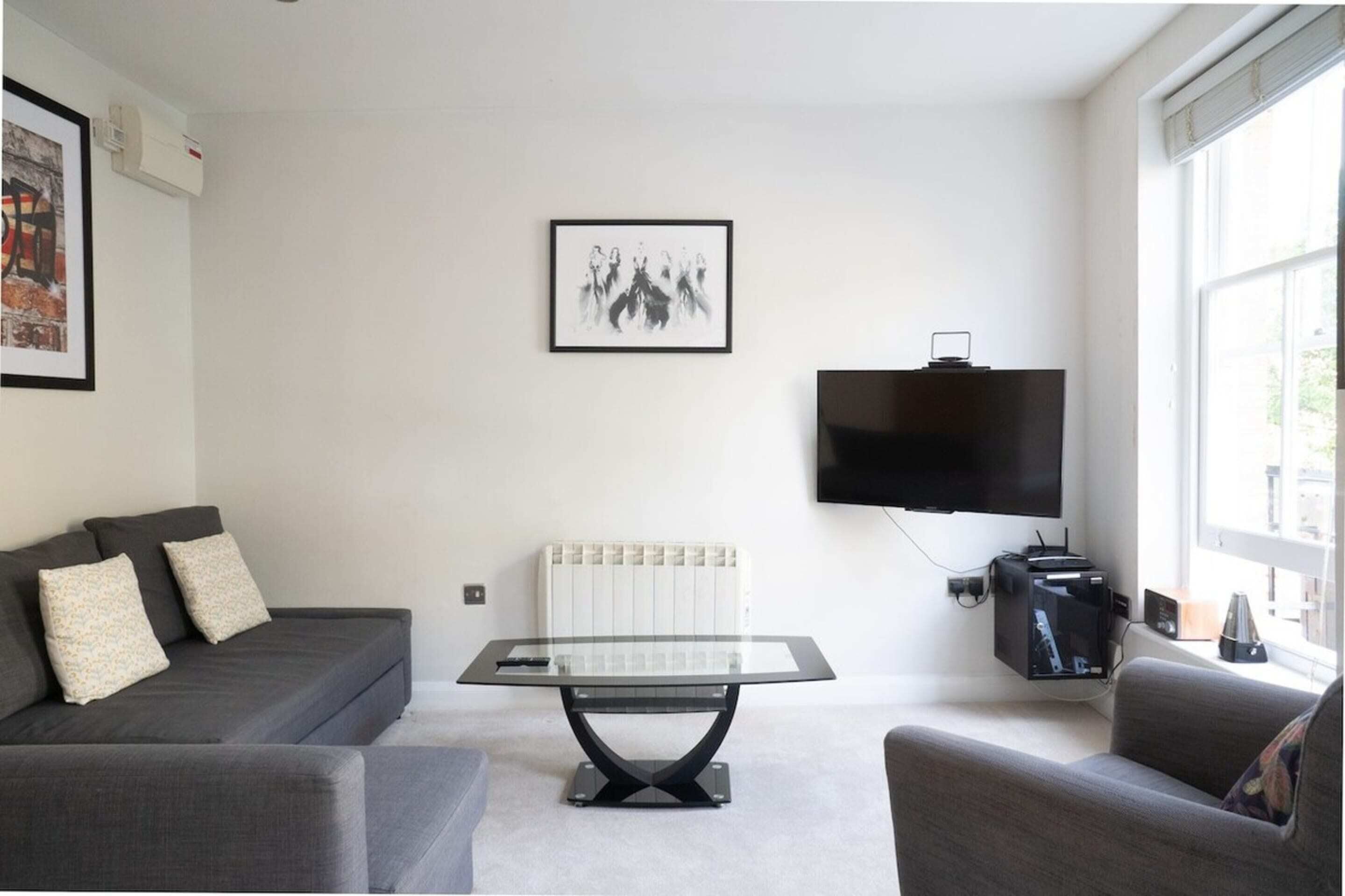 Un salon moderne avec un canapé gris, une table basse en verre, un téléviseur à écran plat et un radiateur.