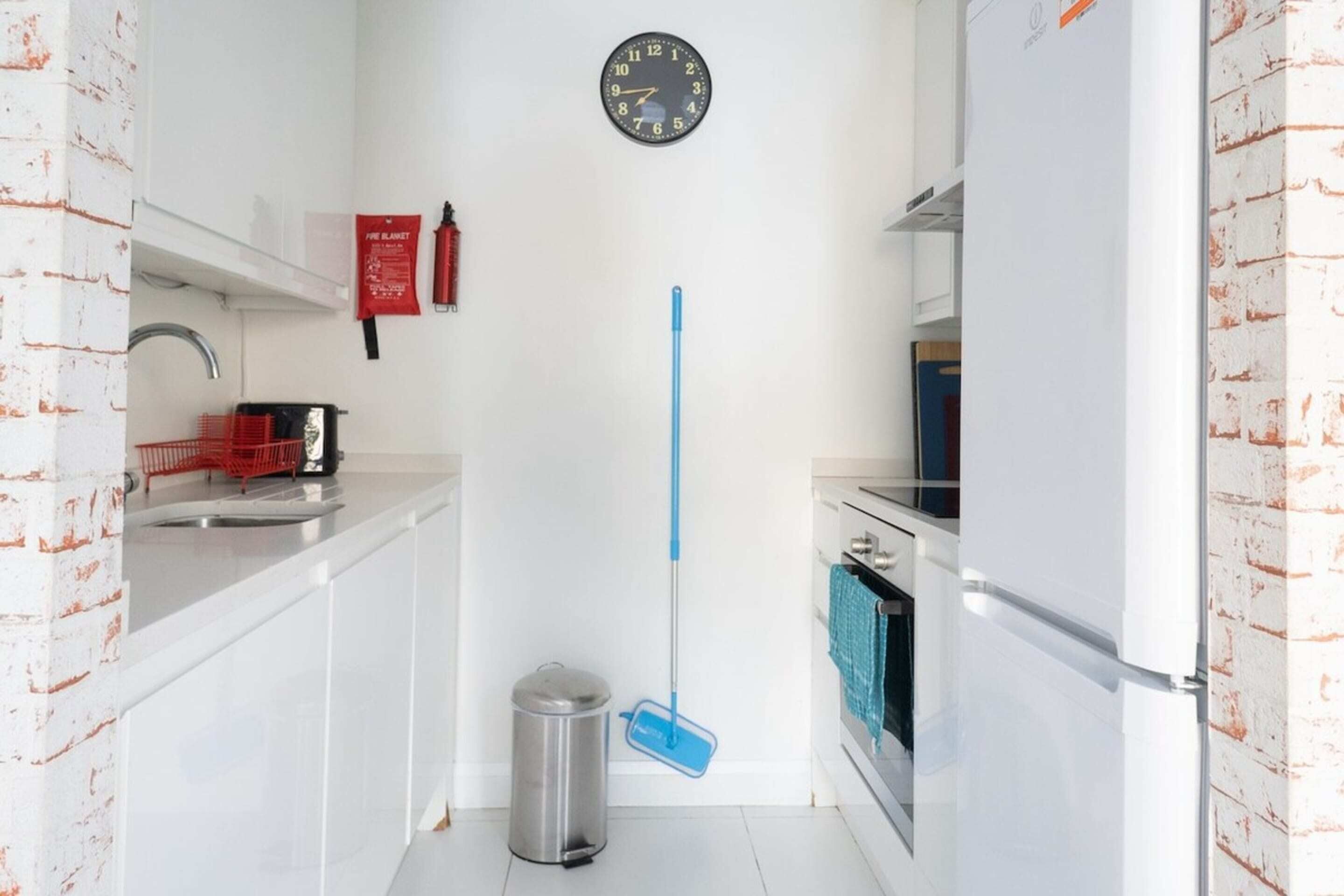 Une cuisine de taille réduite avec des meubles blancs, une poubelle en acier inoxydable, une serpillière bleue, une horloge et une couverture anti-feu accrochée au mur.