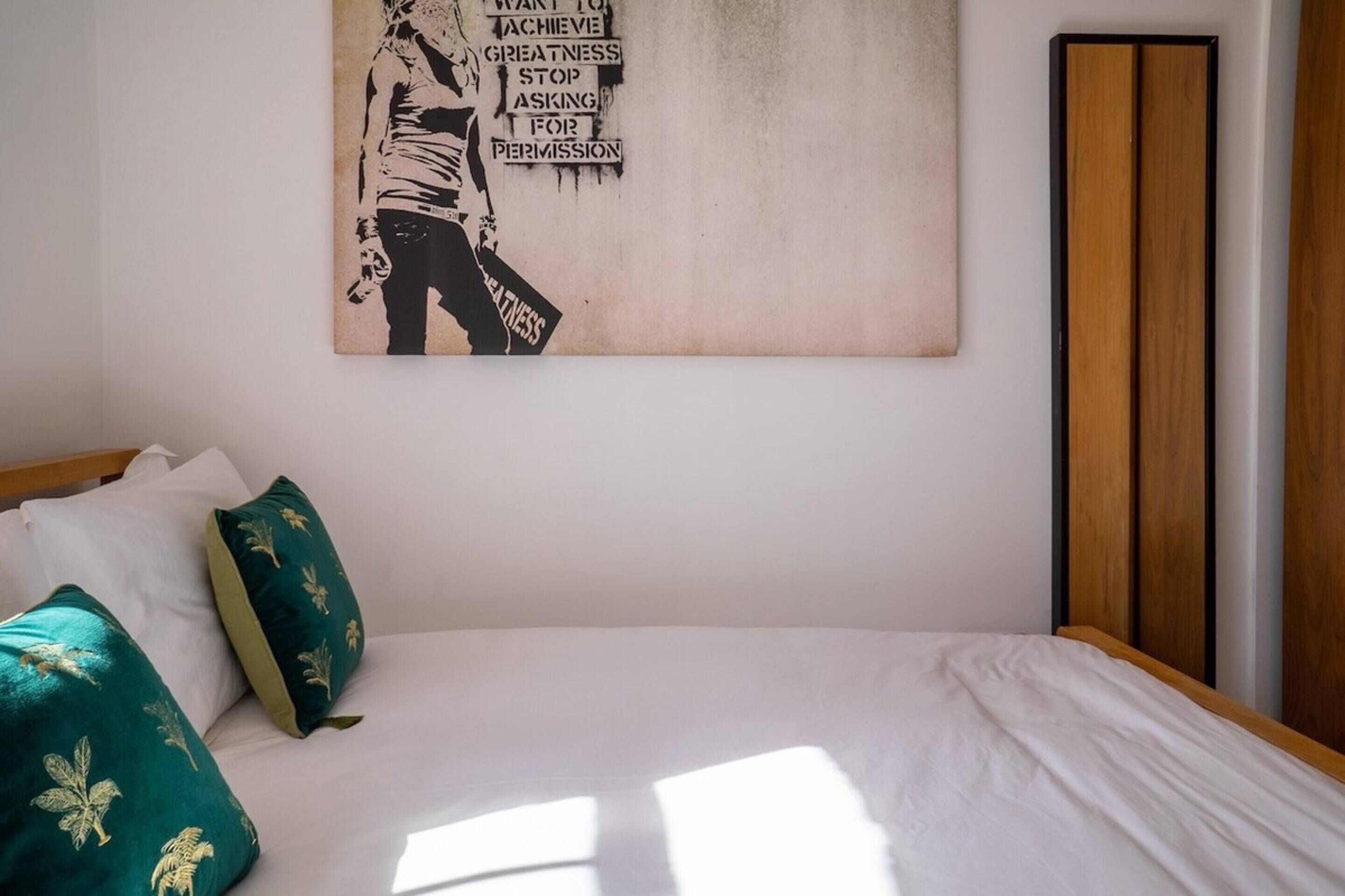 Ein ordentlich bezogenes Bett mit weißen Leinen und zwei grünen Kissen mit Goldblattdekor. Ein an der Wand hängendes Kunstwerk mit einem Schwarz-Weiß-Bild und Text ist sichtbar.