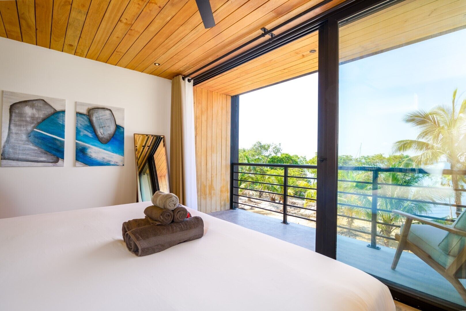 A bedroom with a large bed, two abstract paintings, a mirror, and a balcony with a view of palm trees and greenery.