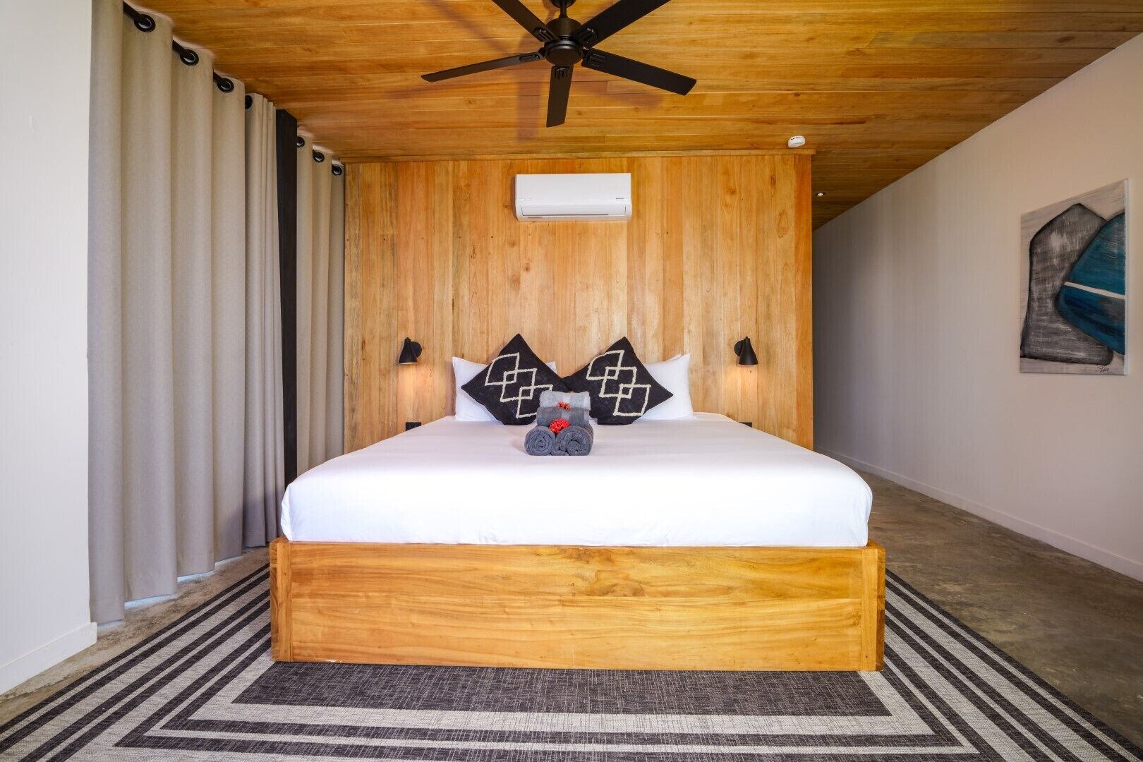 Una camera da letto con una testiera in legno, un letto con biancheria bianca, due cuscini a motivi bianco e neri, un asciugamano arrotolato sul letto, una ventola a soffitto e un quadro appeso al muro.