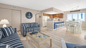 Ein geräumiges Wohnzimmer mit einem gestreiften Sofa, einem Couchtisch und einem Essbereich mit weißen Stühlen sowie einer Küche im Hintergrund.