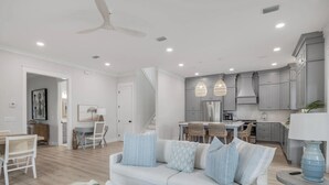 Uma sala de estar moderna com sofá branco, área de jantar e cozinha com armários cinza e luminárias pendentes.