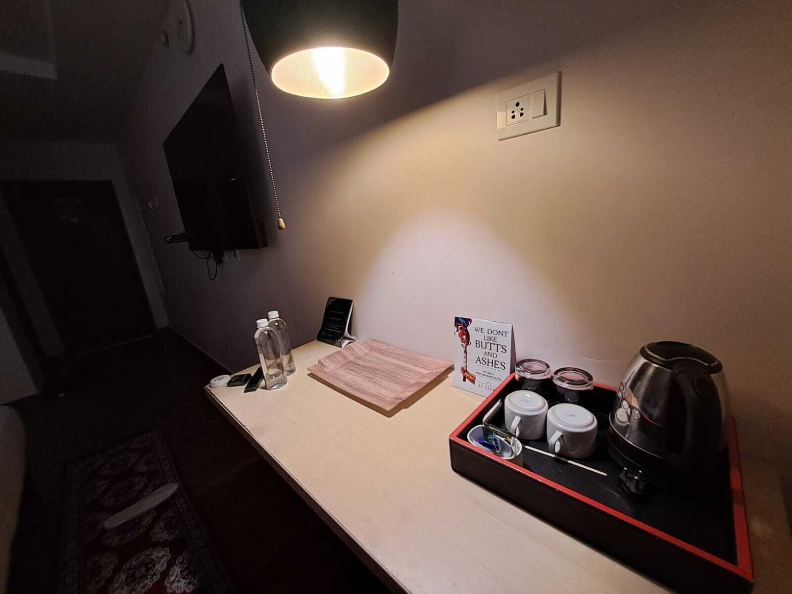 A hotel room with a desk, a lamp, a sign, a tray with mugs and a coffee pot, and a TV mounted on the wall.