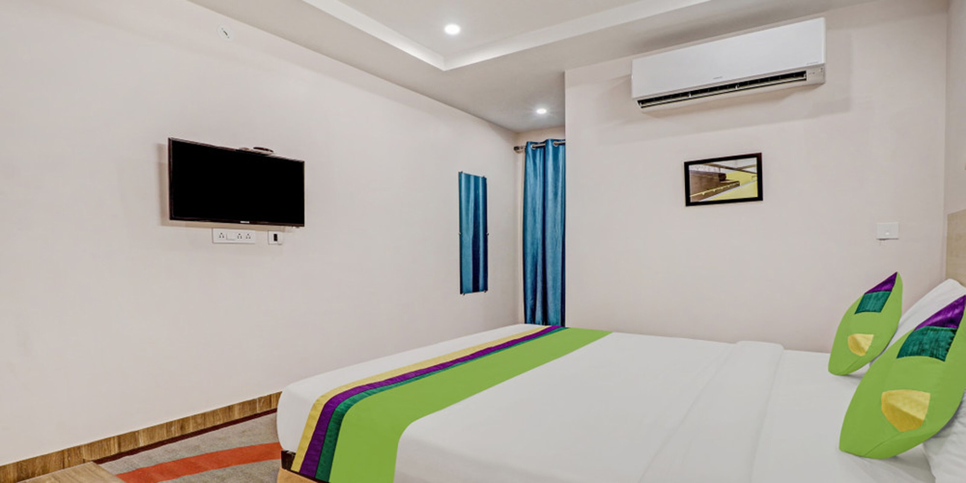 A hotel room with a bed, an air conditioner, a flat-screen TV, and a framed picture on the wall.