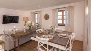 A dining area with a wooden table set for four, white chairs, a TV mounted on the wall, and a sofa in the background. - MY STAY: Relaxation between Chiemsee beach & Kampenwand, plenty of space with 79 sqm (Bernau am Chiemsee)