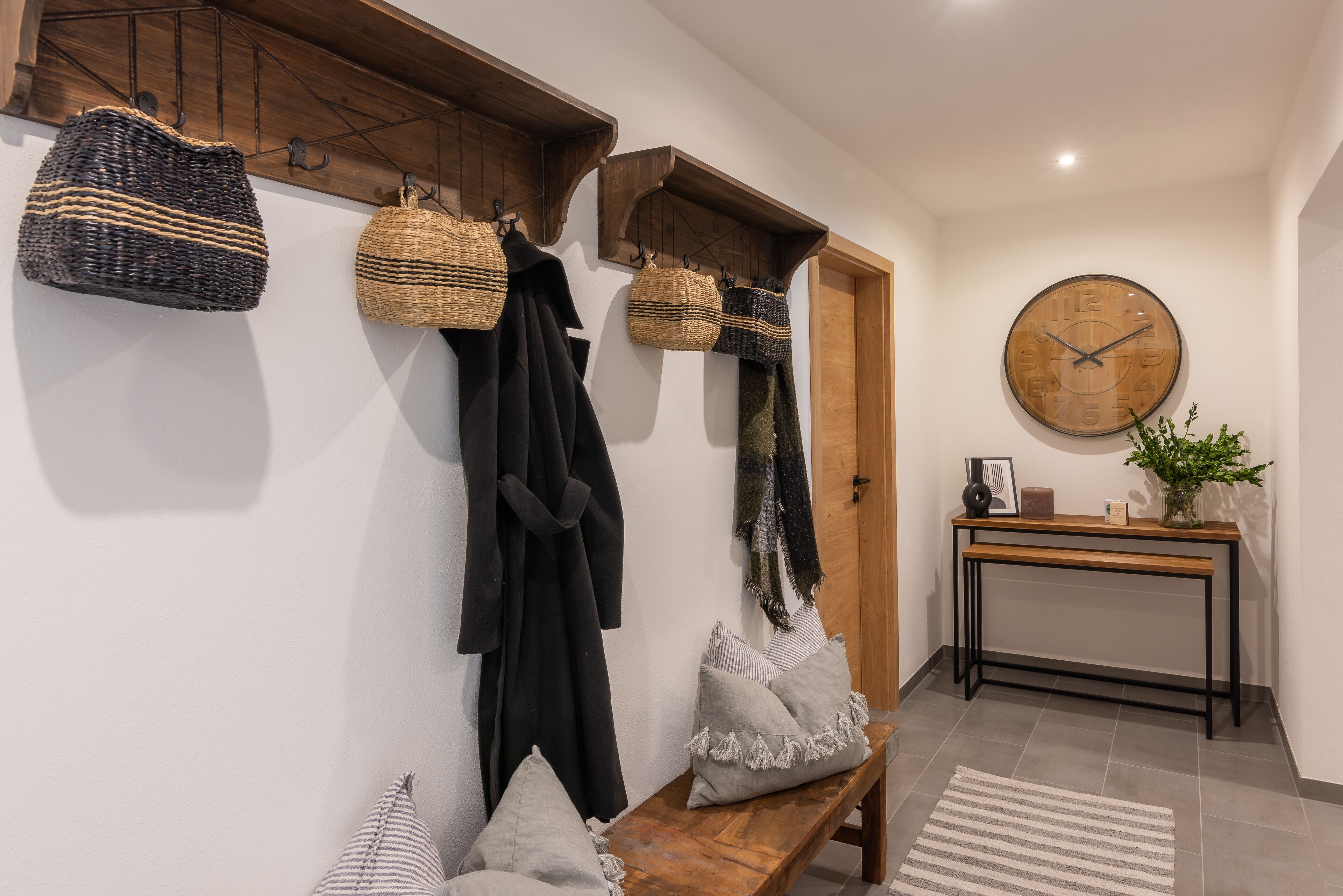A hallway with a coat rack, woven baskets, a bench with pillows, a round wall clock, and a console table with a plant.