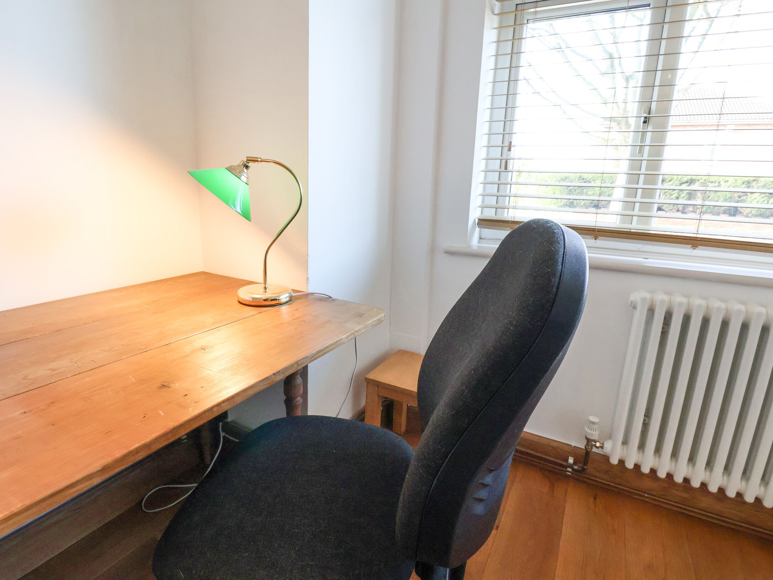 A wooden desk with a green lamp, a black office chair, and a window with blinds.