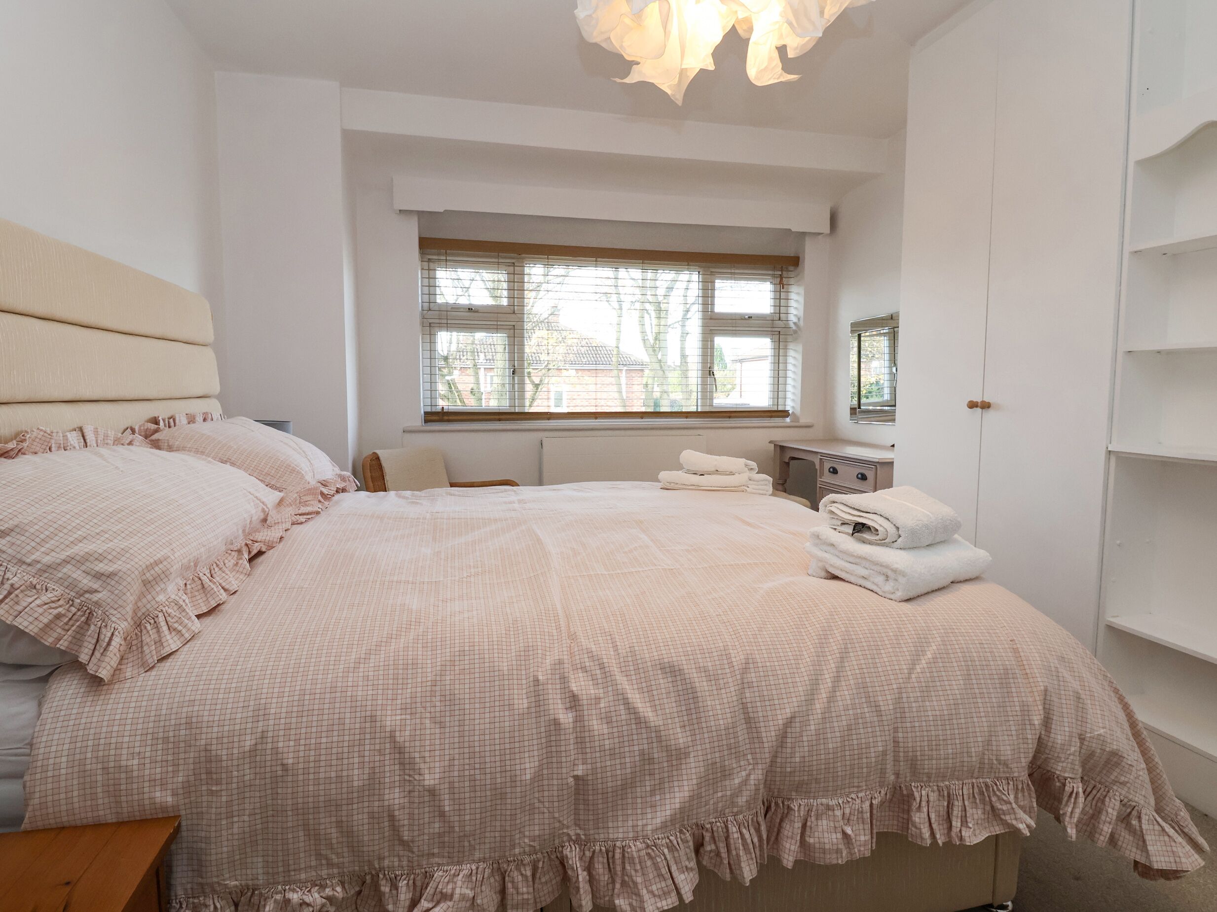A bedroom with a large bed, a bedside table, a window with blinds, and a wardrobe.