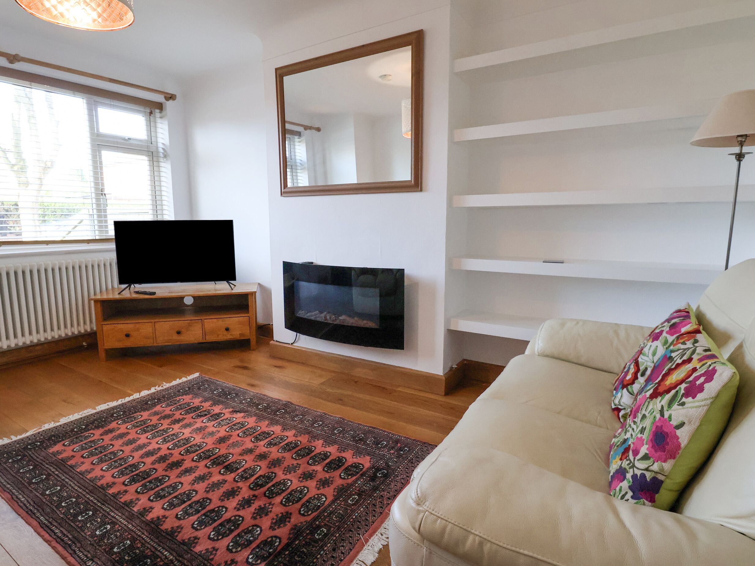 Un salon avec un téléviseur à écran plat posé sur un meuble en bois, un grand miroir au mur, un canapé agrémenté d’un coussin à motifs floraux et un tapis à motifs.