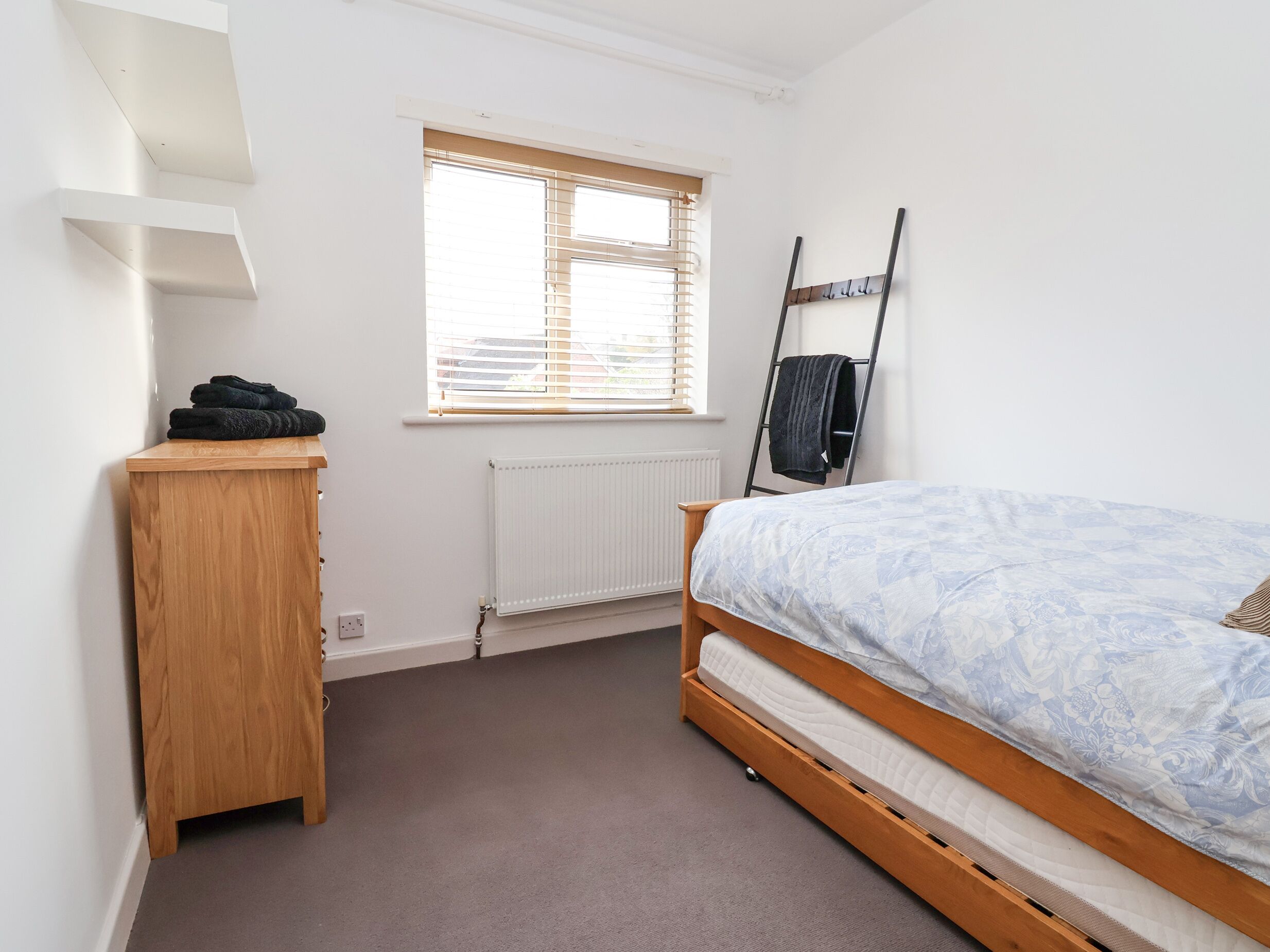 Ein Schlafzimmer mit einem Bett, einer Holzkommode, einem Heizkörper, einem Fenster mit Jalousien und einer an der Wand angelehnten Leiter.