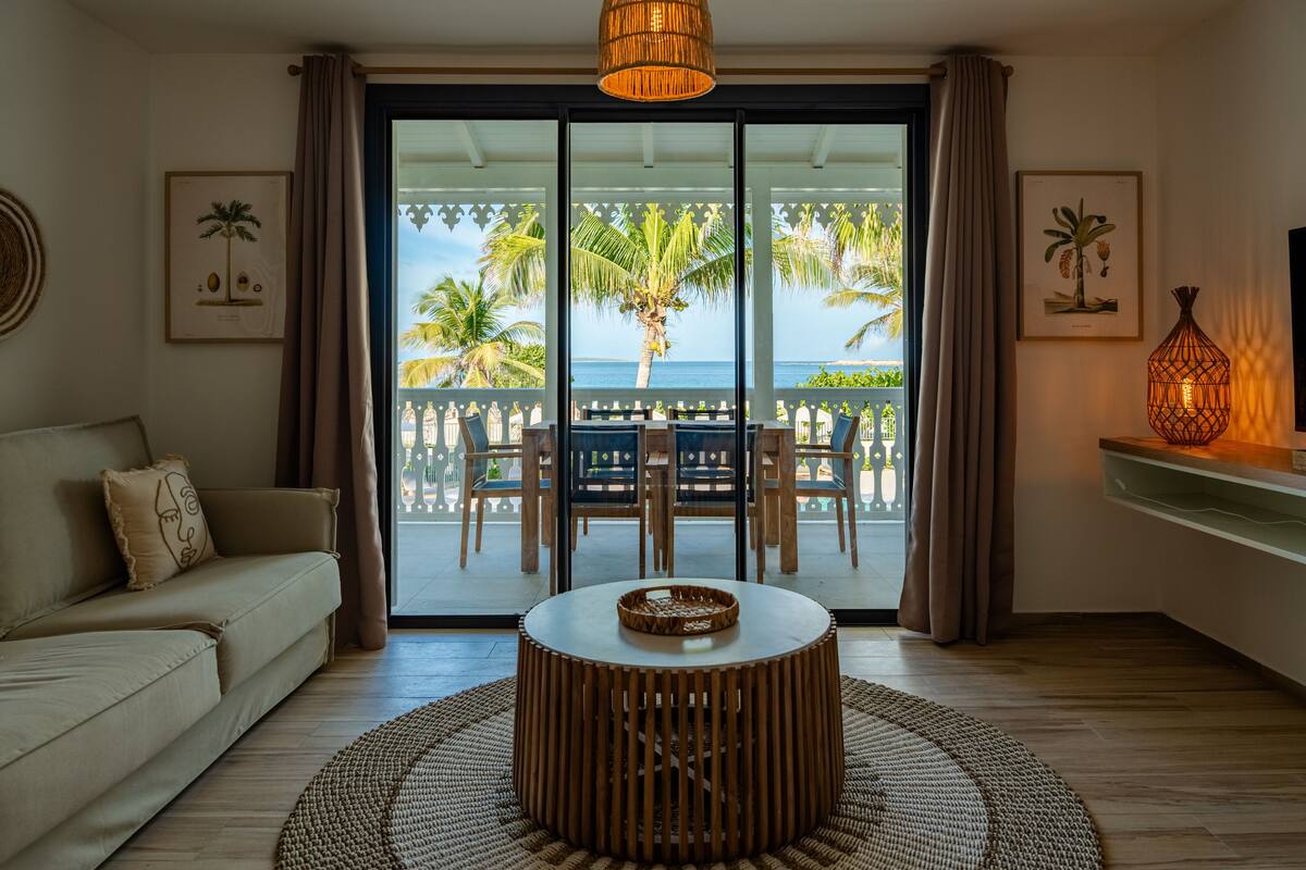 A living room with a sofa, a round wooden coffee table, and a view of a balcony with outdoor furniture and palm trees.
