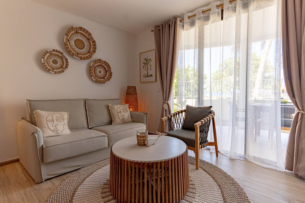 A living room with a beige sofa, a round wooden coffee table, and a patterned rug. There are wall art pieces and a lamp.