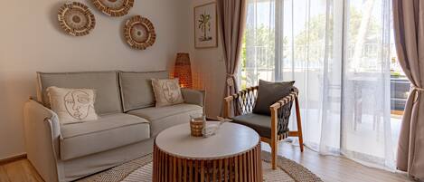 A living room with a beige sofa, a round wooden coffee table, and a patterned rug. There are wall art pieces and a lamp.