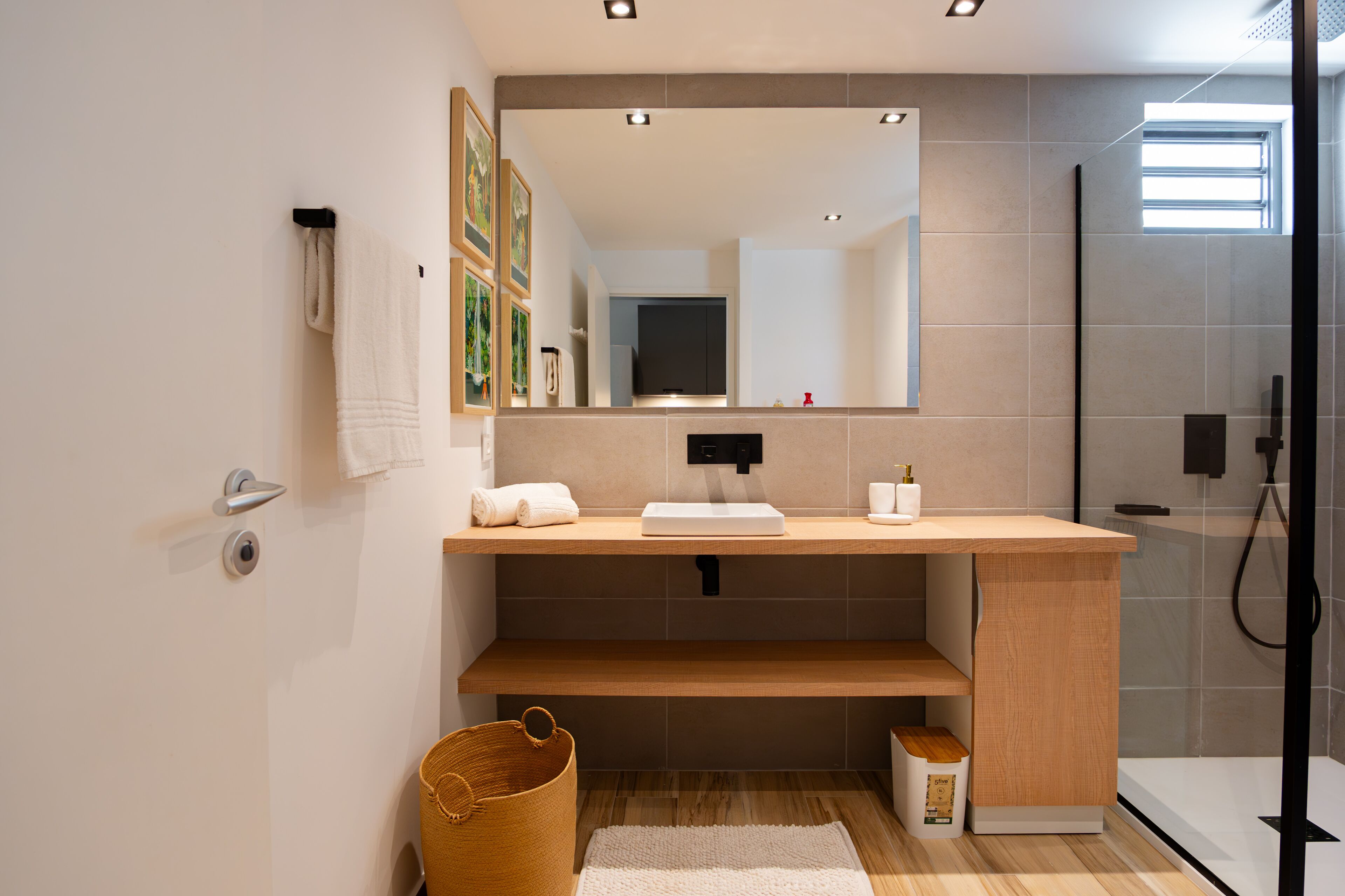 A modern bathroom with a wooden vanity, a large mirror, a shower, and a small trash bin.