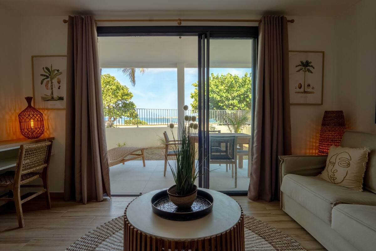 A living room with a sofa, a coffee table with a plant, a wicker chair, a lamp, and a view of the outdoors through a glass door.