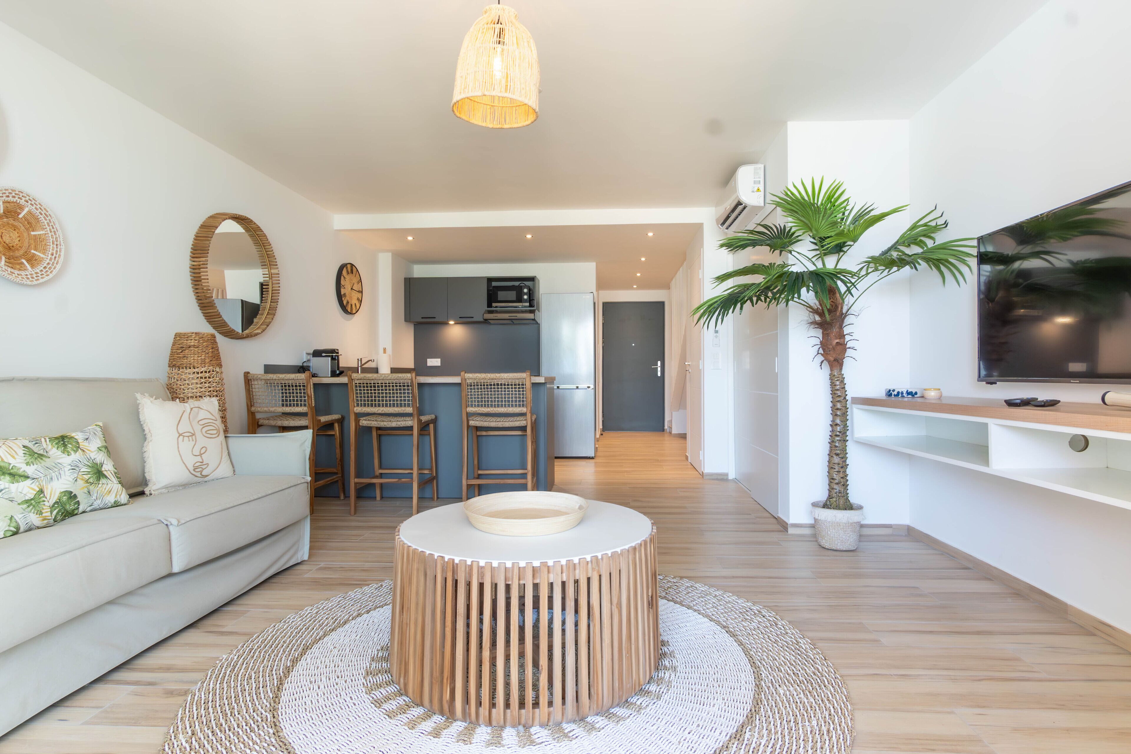 Un salon moderne avec une table basse ronde en bois, un canapé avec des coussins, un tapis tissé et un téléviseur posé sur un meuble.