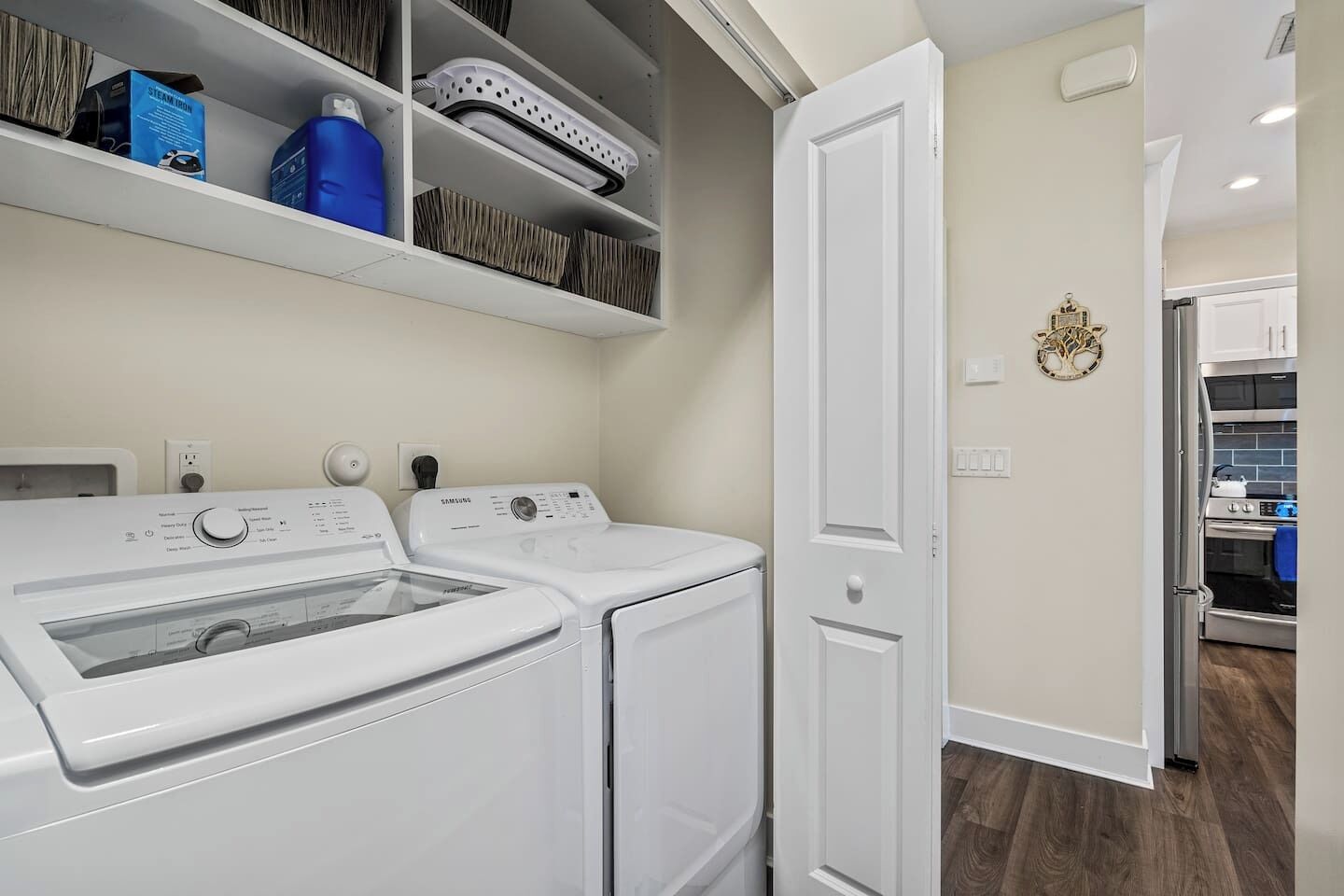 A laundry room with two white front-loading washers and dryers, a shelf with various items, and a door leading to another room.