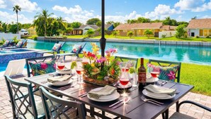 A dining table with chairs, a red umbrella, and a poolside setting with a view of houses and greenery. - Waterfront Htd Pool, Spa, Billiards, Lanai, Canal (Delray Beach)