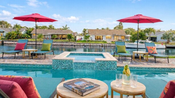 A pool area with lounge chairs, red umbrellas, and a book on a table. - Waterfront Htd Pool, Spa, Billiards, Lanai, Canal (Delray Beach)