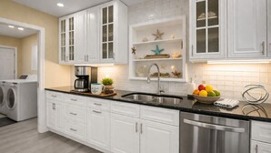 A modern kitchen with white cabinetry, a stainless steel dishwasher, a coffee maker, and a fruit bowl. - Waterfront Htd Pool, Spa, Billiards, Lanai, Canal (Delray Beach)