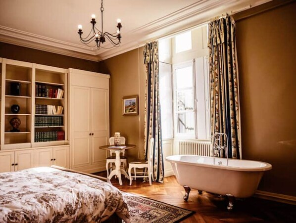 A bedroom with a large bed, a vanity with a bathtub, a small table with a chair, and a bookshelf.