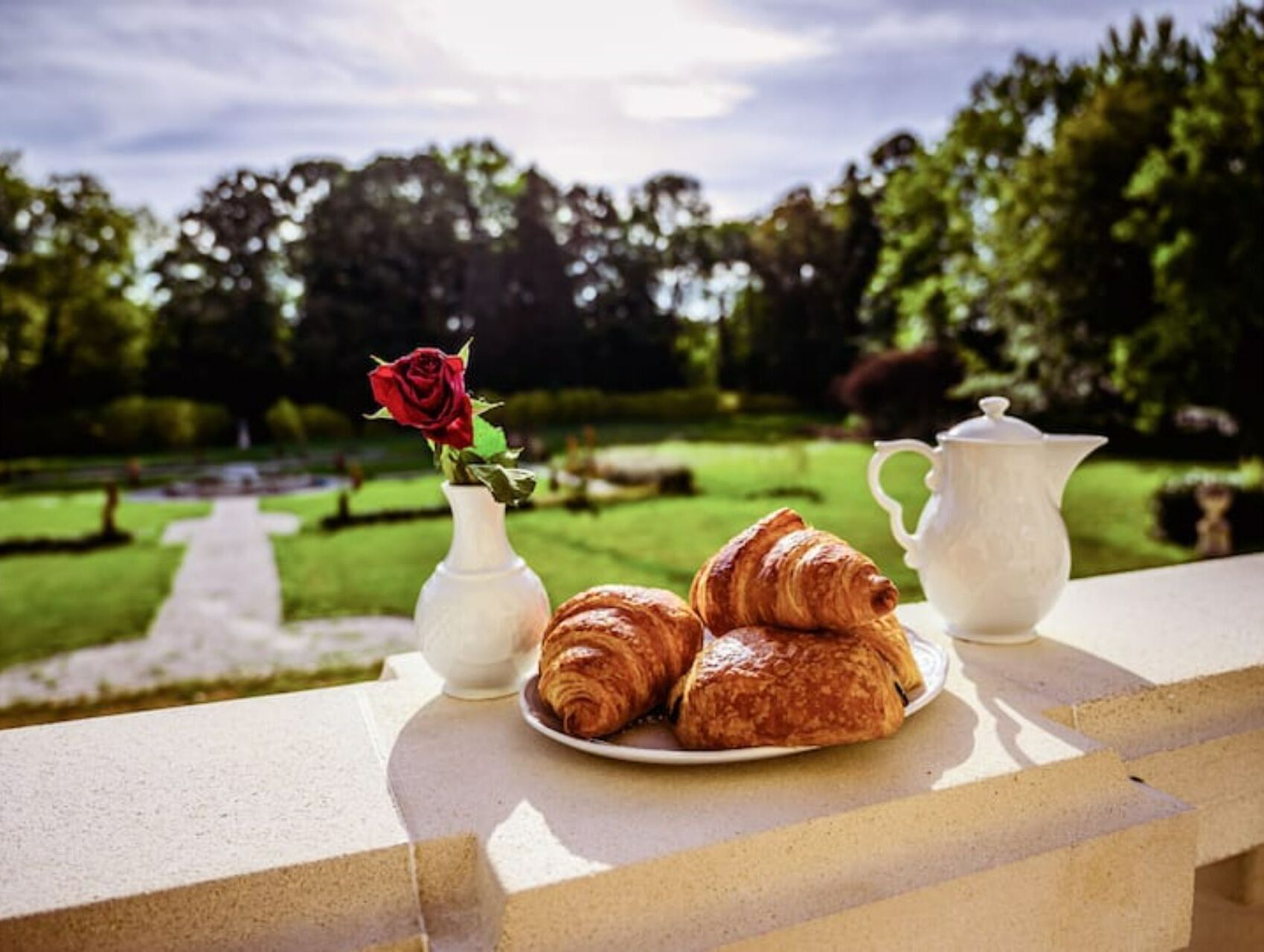 A table with croissants and a teapot on a balcony overlooking a garden.