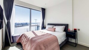Une chambre d’hôtel moderne, dotée d’un grand lit, d’une table de chevet et offrant une vue sur le paysage urbain grâce à de grandes fenêtres.