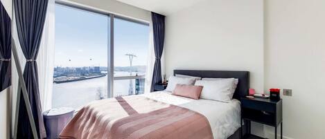 A modern hotel room with a large bed, a nightstand, and a view of a cityscape through large windows.