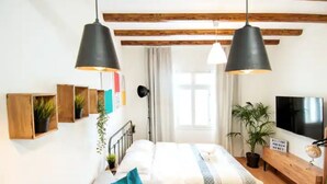 A bedroom with a bed, two hanging black pendant lights, a wooden bench, a TV, and a welcome sign.