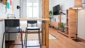 A modern living space with a wooden dining table, two black chairs, a sofa, a TV mounted on the wall, and a lamp.